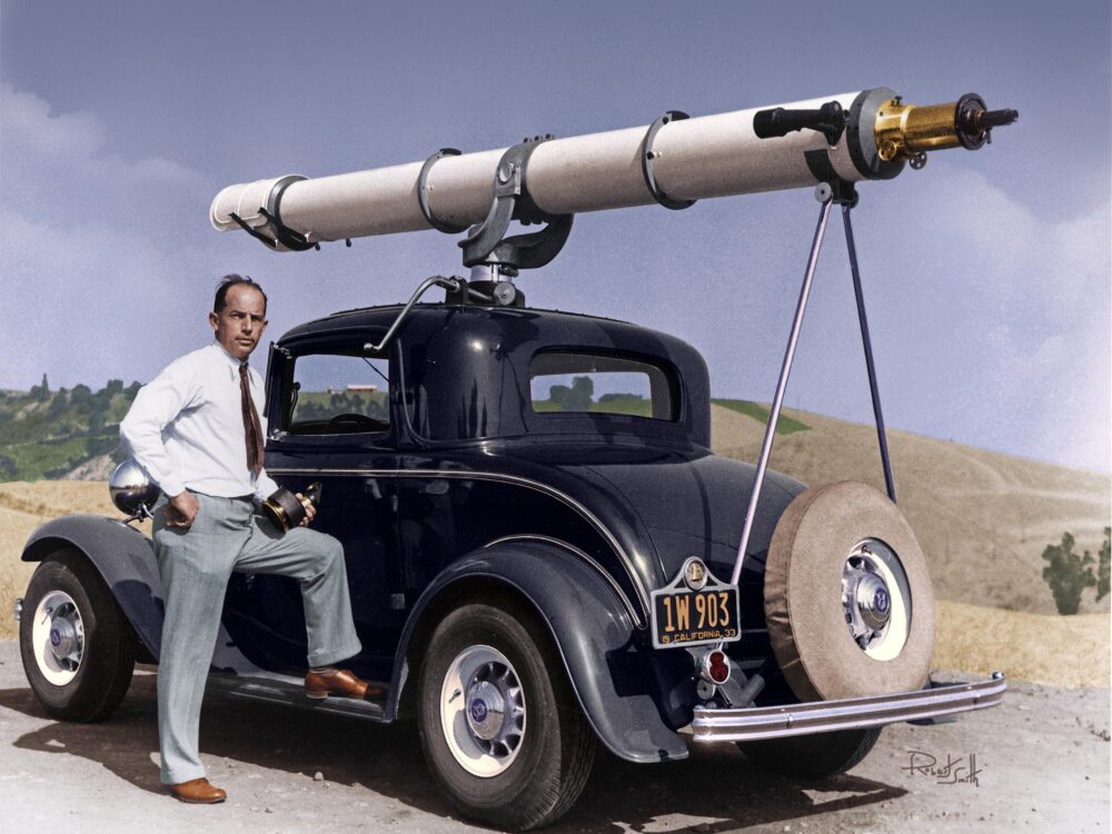 Vintage photo of a man posing in front of a telescope strapped to the hood of a car.
